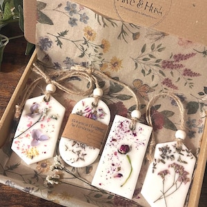 May include: A gift box containing four botanical hanging air fresheners. Each white freshener is decorated with dried flowers and petals, and has a different shape. The box is lined with floral paper and has the text "Hive & Husk" printed on it.