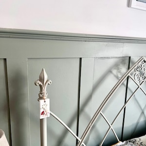 May include: A section of a bed frame with a decorative headboard. The headboard is painted a muted green and features a white metal frame with an ornate design. A tag with floral imagery hangs from the bedpost.