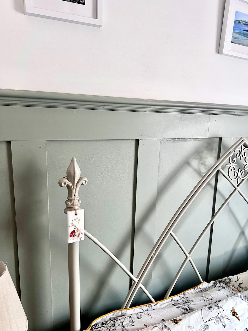 May include: A section of a bed frame with a decorative headboard. The headboard is painted a muted green and features a white metal frame with an ornate design. A tag with floral imagery hangs from the bedpost.