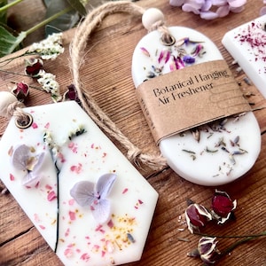 May include: White botanical hanging air fresheners with dried flowers and a twine loop for hanging. One has a brown paper label that reads "Botanical Hanging Air Freshener". The air fresheners are on a wooden surface.