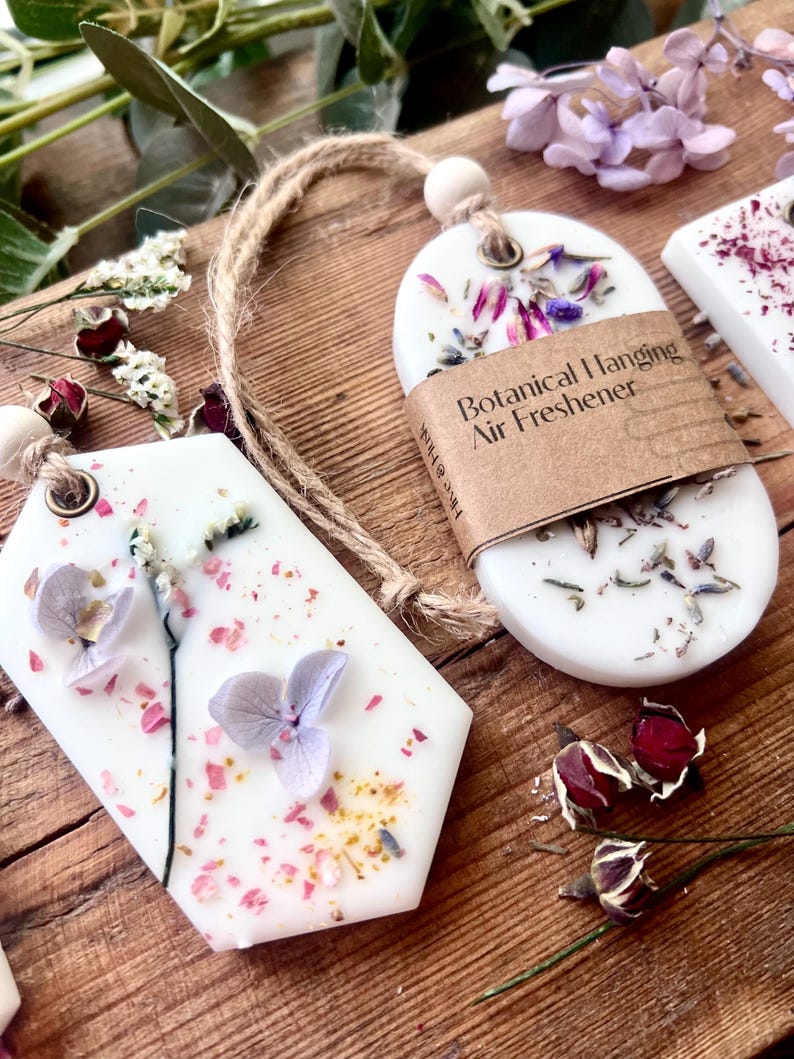 May include: White botanical hanging air fresheners with dried flowers and a twine loop for hanging. One has a brown paper label that reads "Botanical Hanging Air Freshener". The air fresheners are on a wooden surface.