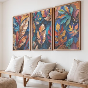 Op de afbeelding: Drie ingelijste kunstwerken met kleurrijke bladeren in rood, oranje, blauw en geel. De kunst hangt boven een houten bank met witte kussens en een gevlochten mand, tegen een witte muur.
