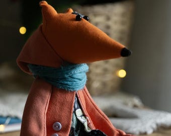 May include: A plush fox toy wearing a coral colored jacket with buttons and a blue scarf. The fox has black eyes and a long orange snout.