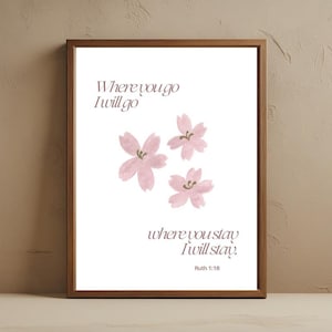 May include: A framed print with a white background features the text "Where you go I will go, where you stay I will stay. Ruth 1:16." Three pink watercolor flowers are in the center. The frame is a dark brown color.