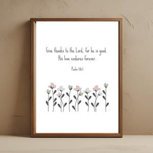 Pode incluir: Uma impressão emoldurada com fundo branco apresenta o texto "Dai graças ao Senhor, porque ele é bom. O seu amor dura para sempre. Salmo 136:1." Ilustrações florais em rosa, branco e verde adornam a parte inferior. A moldura é marrom quente.