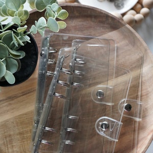 May include: A set of three clear plastic binders with silver-toned metal ring mechanisms and snap button closures. Each binder features a transparent cover, allowing the contents to be visible, and is designed for organising loose-leaf paper.