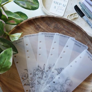 May include: A wooden tray displays several translucent planner inserts with floral line art and text. The inserts are labelled with phrases like "High Priority Savings" and "Cash Denomination Savings." A small card with the text "One Step at a Time" is also visible.