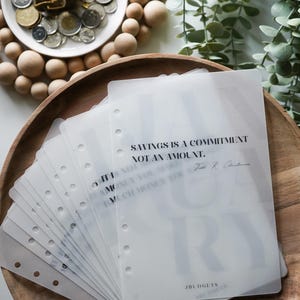 Può includere: Una pila di fogli di plastica bianca traslucida con fori per un raccoglitore. Il foglio superiore ha il testo "SAVINGS IS A COMMITMENT NOT AN AMOUNT." e una firma. Il foglio inferiore ha il testo "JBUDGETS".