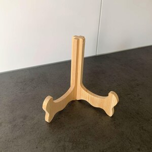 May include: A light-coloured wooden display stand with a three-legged design. The stand is made of unfinished wood and is designed to hold a plate or other decorative item. The stand is approximately 20 cm tall.