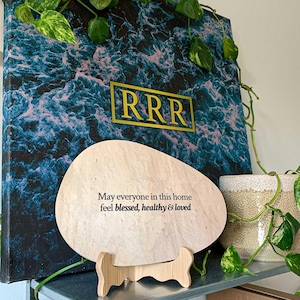 May include: A wooden sign with the text "May everyone in this home feel blessed, healthy & loved" on a wooden stand. A canvas with the letters "RRR" in a yellow rectangle is in the background. Green vines and a decorative pot are also visible.
