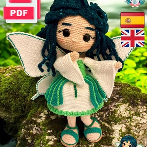 Puede incluir: Muñeca de hada de ganchillo con alas blancas, un vestido verde y blanco y cabello verde azulado. La muñeca tiene ojos negros grandes y usa sandalias verdes. Un icono PDF está en la esquina superior izquierda.