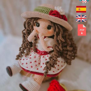 May include: A crocheted doll wearing a white dress with red polka dots, a brown hat with a green band and red flowers, and brown curly hair. The doll is holding a red rose.