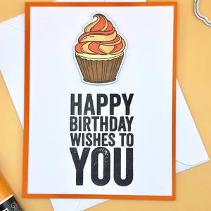 Könnte beinhalten: Geburtstagskarte mit weißem Hintergrund und orangefarbenem Rand. Die Karte zeigt eine Cupcake-Illustration und den Text "HAPPY BIRTHDAY WISHES TO YOU" in fetten schwarzen Buchstaben. Ein weißer Umschlag befindet sich hinter der Karte.