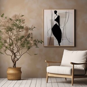 May include: An abstract art print featuring a black figure on a white background, framed in a light wood. A potted plant with a beige pot and a white armchair with wooden frame complete the interior scene. The wall is painted in a warm, neutral tone.