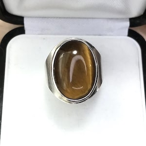 Puede incluir: Un anillo de plata con una gran piedra ojo de tigre ovalada, exhibido en una caja de anillos en blanco y negro. El anillo tiene una banda de plata pulida. Otras cajas de joyería vintage están en el fondo.