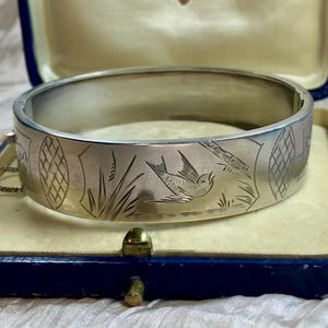 Puede incluir: Una pulsera brazalete plateada con bisagras con diseños grabados de pájaros y geométricos, presentada en una caja de joyería azul y dorada. La pulsera presenta un pájaro detallado, follaje y patrones en forma de diamante. La caja está abierta, revelando un interior crema.