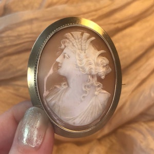 Può includere: Un broche cammeo con un cammeo bianco a profilo di donna in una cornice ovale d'oro.