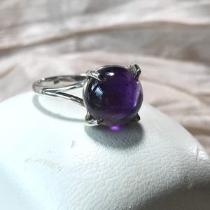 以下が含まれることがあります： 大きな丸い紫色の紫水晶の宝石をあしらったシルバーリング。宝石は4つの爪で固定されています。リングは、柔らかくニュートラルな色調の背景を持つ白い表面にセットされています。