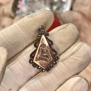 May include: A vintage pendant with a deer design. The pendant features a copper-colored diamond-shaped center with a deer silhouette, framed by a silver-toned border with small, dark gemstones. The pendant is suspended from a silver bail.