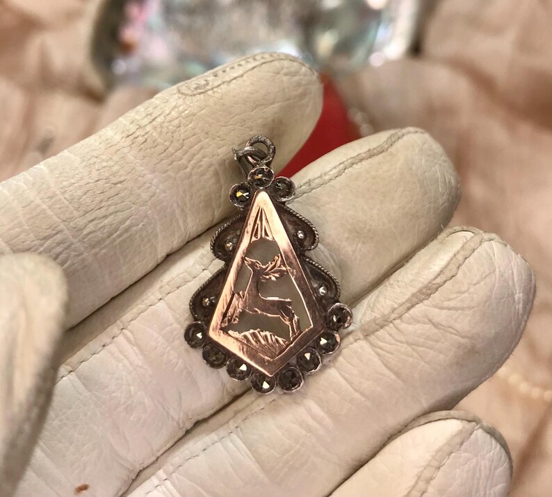 May include: A vintage pendant with a deer design. The pendant features a copper-colored diamond-shaped center with a deer silhouette, framed by a silver-toned border with small, dark gemstones. The pendant is suspended from a silver bail.