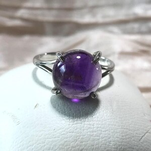 May include: A silver ring with a large, round, polished amethyst gemstone. The deep purple stone is held by four prongs. The ring is set against a white surface and a soft, neutral background.