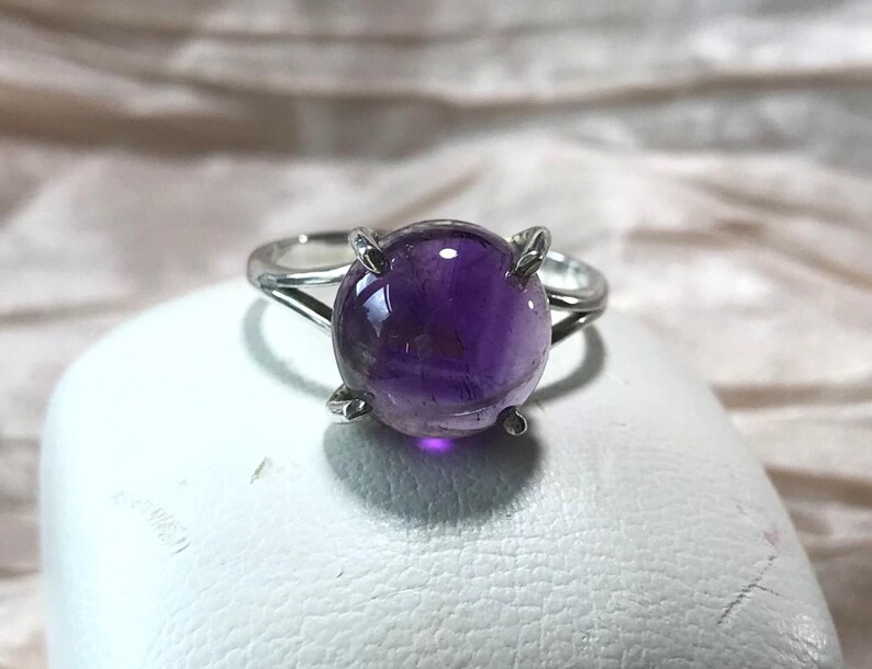 May include: A silver ring with a large, round, polished amethyst gemstone. The deep purple stone is held by four prongs. The ring is set against a white surface and a soft, neutral background.