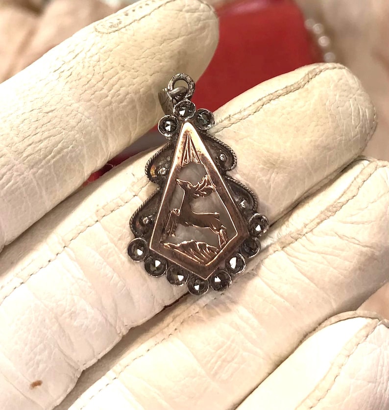 May include: A vintage silver pendant with a deer silhouette inside a diamond-shaped frame. The pendant has a decorative border with small, dark stones and a loop for a chain. The background is a white surface.