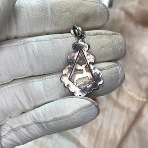 May include: Silver pendant with a deer silhouette inside a triangular frame. The pendant has a decorative, scalloped edge and a loop for a chain. The pendant is held in a white glove.