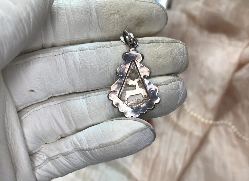 May include: Silver pendant with a deer silhouette inside a triangular frame. The pendant has a decorative, scalloped edge and a loop for a chain. The pendant is held in a white glove.