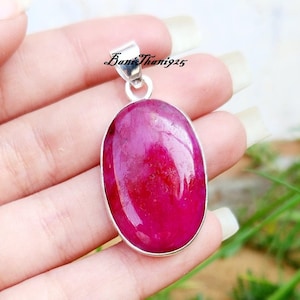 Peut inclure: Un pendentif en argent avec une grande pierre précieuse ovale de couleur rose foncé. Le pendentif est tenu dans une main sur un fond vert.