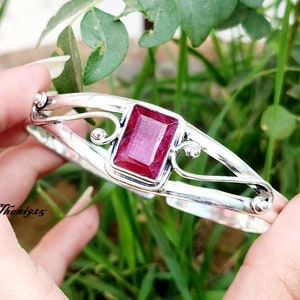 May include: A silver bangle bracelet with a large, rectangular, red gemstone set in the center. The bracelet has a delicate, ornate design.