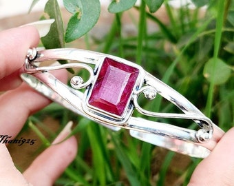 May include: A silver bangle bracelet featuring a large, rectangular, red gemstone set in the centre. The bracelet has a delicate, ornate design.