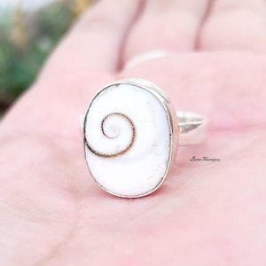 Puede incluir: Un anillo de plata con una concha ovalada blanca con un patrón de espiral marrón.