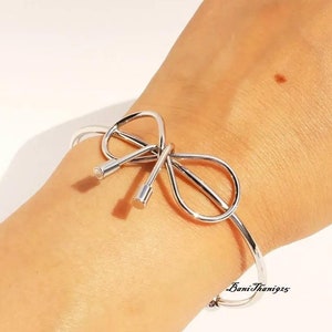 Puede incluir: Un brazalete de plata con un diseño de lazo. El brazalete está abierto y tiene un aspecto simple y elegante.
