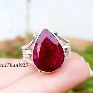 Peut inclure: Bague en argent ornée d'une grosse pierre rouge foncé en forme de poire. La bague a un anneau en argent détaillé avec des éléments décoratifs. Le texte "BaniThani925" est visible. La bague est présentée sur un fond naturel flou.