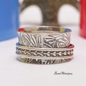 May include: A silver spinning ring with a textured design. The ring has three bands, the top band has a leaf pattern, the middle band has a braided pattern, and the bottom band has a textured pattern.