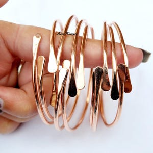 Puede incluir: Conjunto de seis brazaletes de cobre de color oro rosa con un extremo en forma de lágrima. Los brazaletes están apilados uno encima del otro.
