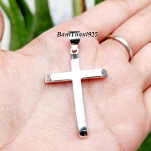 Pode incluir: Pingente de cruz prateada com um design simples e polido. A cruz &eacute; segurada em uma m&atilde;o, com o texto "BaniThani925" vis&iacute;vel. Um s&iacute;mbolo religioso.