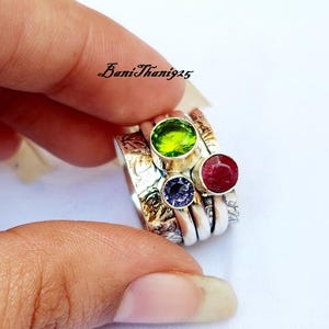 May include: A silver ring with four stacked bands, each with a different colored gemstone. The top band has a green gemstone, the second band has a red gemstone, the third band has a purple gemstone, and the bottom band is plain silver.