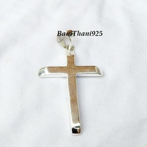 Pode incluir: Um pingente de cruz prateada com bordas arredondadas, suspenso por um pequeno la&ccedil;o. A cruz &eacute; exibida em um fundo branco. O texto "BanThani925" &eacute; vis&iacute;vel na imagem.