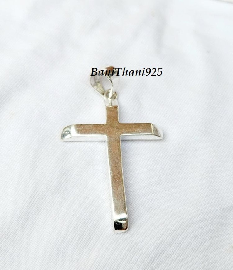 Pode incluir: Um pingente de cruz prateada com bordas arredondadas, suspenso por um pequeno la&ccedil;o. A cruz &eacute; exibida em um fundo branco. O texto "BanThani925" &eacute; vis&iacute;vel na imagem.