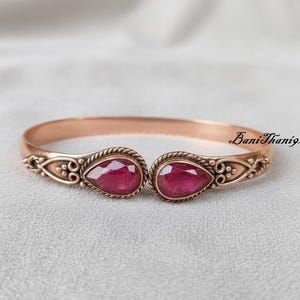 Op de afbeelding: Een roségouden manchetarmband met twee druppelvormige, dieprode edelstenen. De armband heeft sierlijke details rond de edelstenen en de tekst "BaniThani925". De armband wordt gepresenteerd op een lichtgrijze ondergrond.