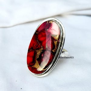 May include: Silver ring featuring an oval-cut stone with a striking red and gold pattern. The stone's design incorporates red, gold, and white colours. This ring is a bold statement piece, suitable for various occasions.