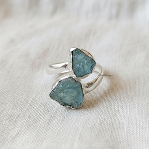 Peut inclure: Une bague en argent ornée de deux pierres d'aigue-marine bleu clair brutes. Les pierres sont serties dans un anneau en argent qui entoure le doigt. La bague est présentée sur une surface blanche texturée.
