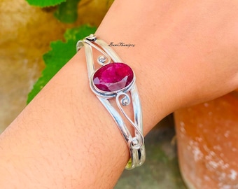 Puede incluir: Un brazalete de plata con una gran piedra preciosa de rubí de forma ovalada engastada en el centro. El brazalete tiene un diseño delicado e intrincado con espirales y curvas.
