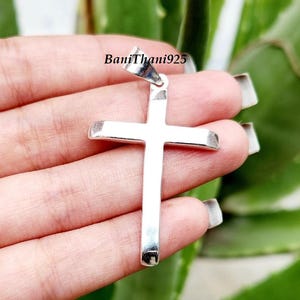 Pode incluir: Um pingente de cruz de prata com acabamento polido. A cruz &eacute; segurada em uma m&atilde;o contra um fundo verde desfocado. O texto "BaniThani925" &eacute; vis&iacute;vel na fian&ccedil;a do pingente.