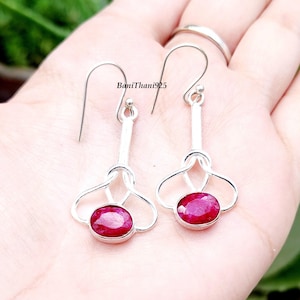 May include: A pair of silver earrings with a drop design. Each earring features a vibrant, oval-shaped ruby gemstone set in a silver bezel. The earrings have a hook closure and a decorative, openwork design. The text "BaniThani925" is visible.