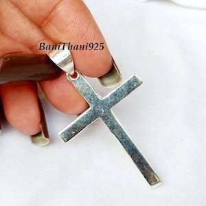 Pode incluir: Um pingente de cruz prateada com uma argola para corrente. A cruz &eacute; segurada por uma m&atilde;o contra um fundo branco. O texto "BaniThani925" &eacute; vis&iacute;vel no pingente.