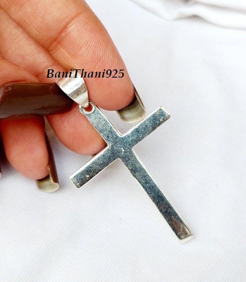 Pode incluir: Um pingente de cruz prateada com uma argola para corrente. A cruz &eacute; segurada por uma m&atilde;o contra um fundo branco. O texto "BaniThani925" &eacute; vis&iacute;vel no pingente.