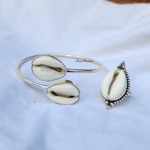May include: A silver bangle bracelet and ring set featuring white cowrie shell accents. The bangle is open and has two cowrie shells attached to it. The ring has a single cowrie shell set in a silver band with a beaded border.